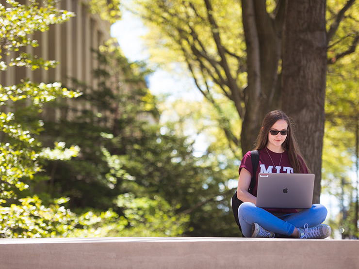 Woman wearing an M-I-T shirt sitting on a wall outside of M-I-T with a laptop open on her lap.
