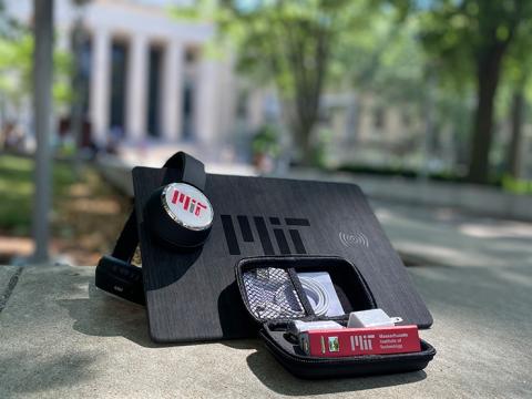 A pair of headphones, a rectangular mouse pad, and a portable phone charger, each with the M-I-T logo on it. They are on a wall with the main entrance to M-I-T at 77 Massachusetts Avenue in the background.