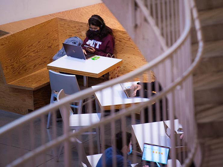 A woman wearing an M-I-T sweatshirt sitting indoors at a table with an open laptop also looking at her phone. Other students are sitting at tables nearby using laptops.