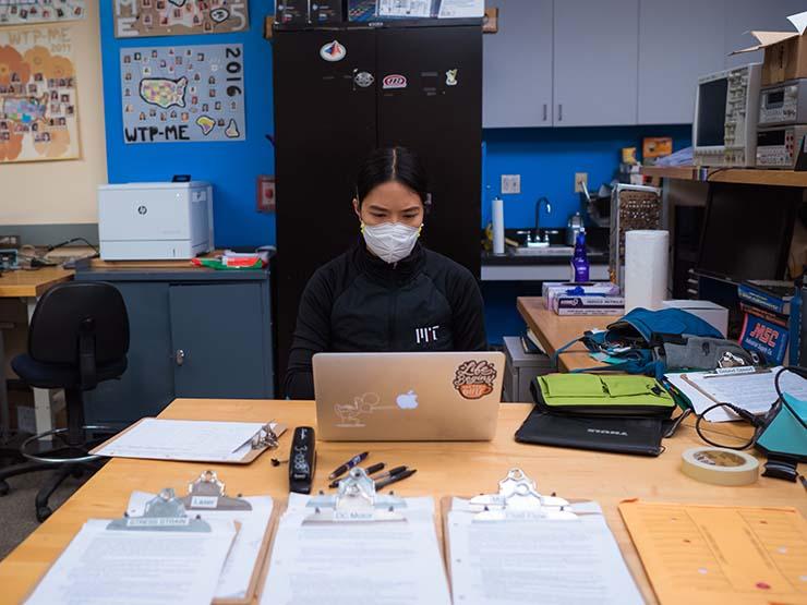 A woman wearing a mask and an M-I-T shirt in a lap looking at her laptop.