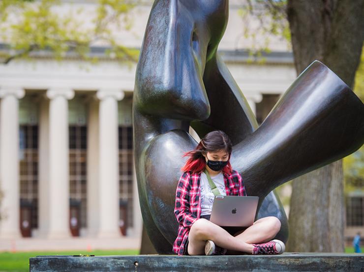 A girl wearing a mask sitting outside of M-I-T using her laptop.