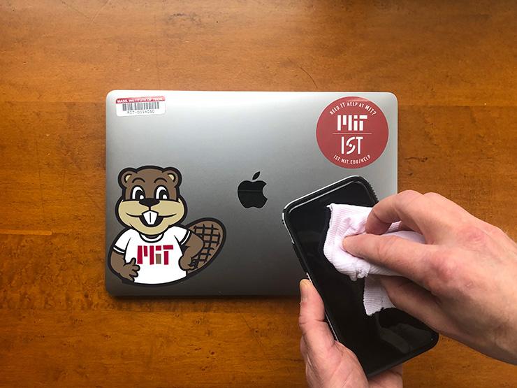 Hands wiping the screen of a smartphone with a cloth. In the background is a closed Apple laptop with M-I-T stickers on it.