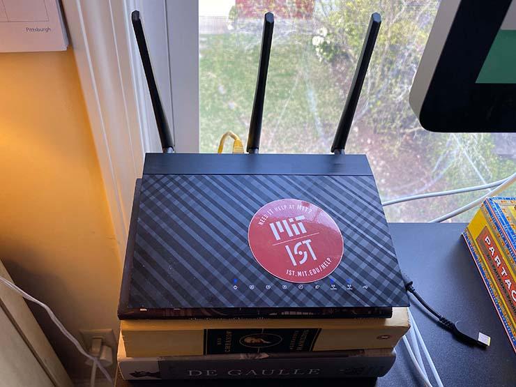 A home computer router on a stack of books next to a computer monitor in front of a window.