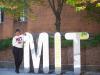 A woman wearing an M-I-T sweatshirt looking at her phone standing outside next to large M I and T letters. On the I are the words The Stata Center.