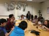 Students eating lunch around a conference room table. Silver balloon letters spelling out “IS&T” are floating against a wall in the background against a whiteboard.